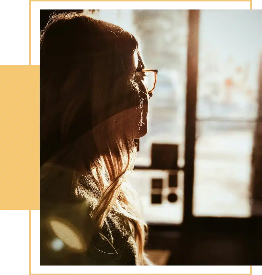 Woman with long hair and glasses looking out a sunlit window.