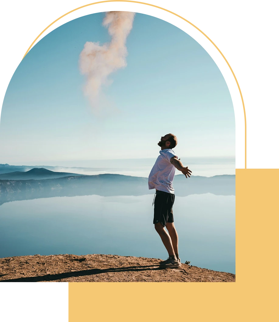 Man standing on a cliff with arms outstretched, facing the sky over a calm lake and mountains.