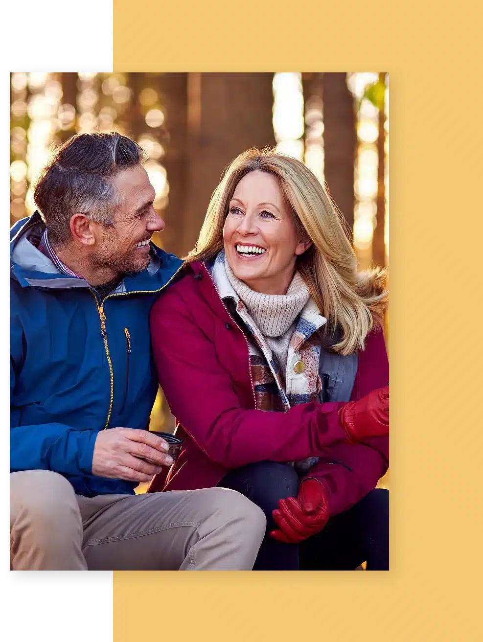 A middle-aged couple in winter coats smiles at each other in a forest.