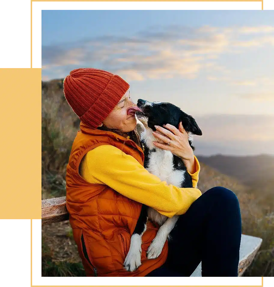 A smiling woman wearing a beanie and vest holds her dog, who is licking her face.